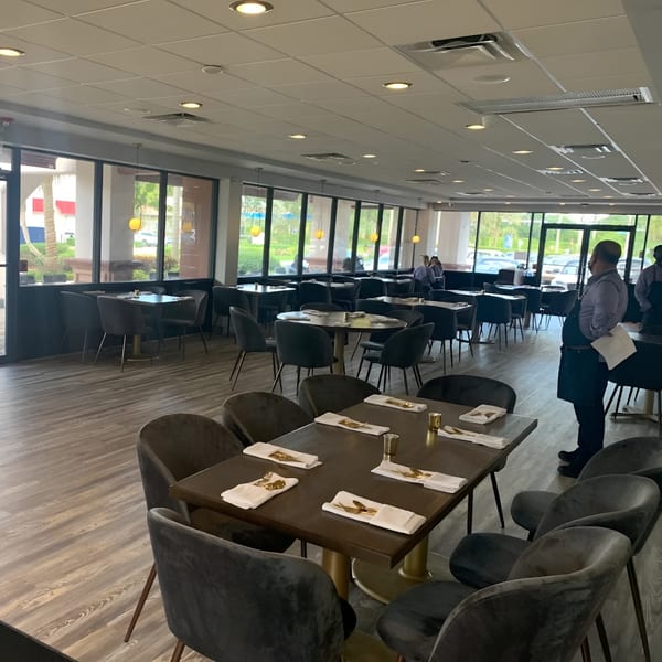 Carmela's dining room with set tables, dark chairs, large windows, and a server standing.