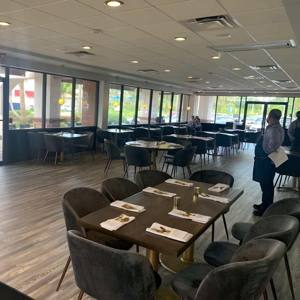 Carmela's dining room with set tables, dark chairs, large windows, and a server standing.
