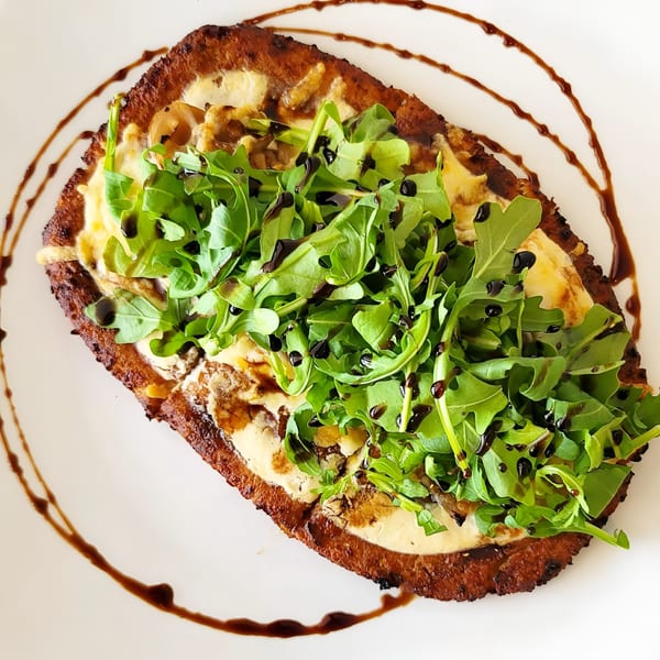 Crispy flatbread topped with creamy sauce, arugula, and balsamic glaze on a white plate.