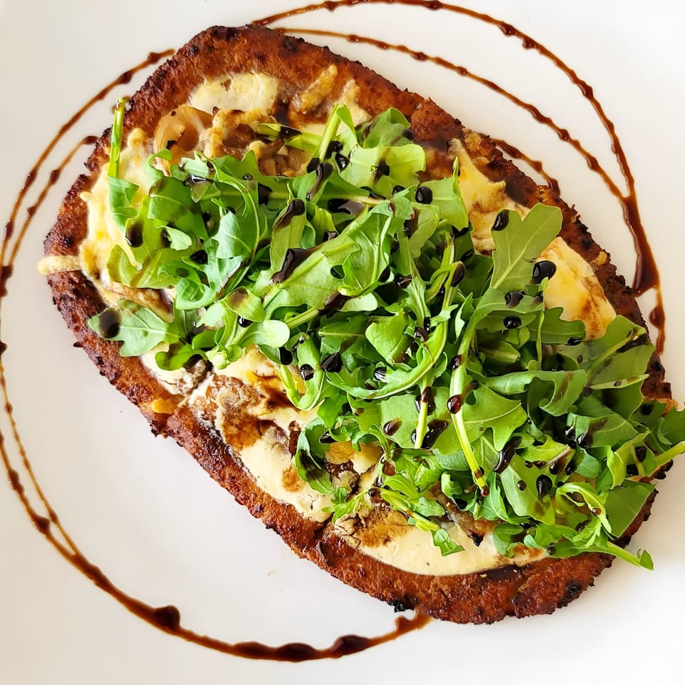 Crispy flatbread topped with creamy sauce, arugula, and balsamic glaze on a white plate.