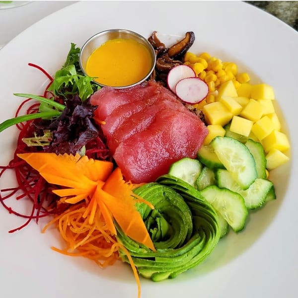 A vibrant poke bowl with tuna, avocado, mango, cucumber, corn, radishes, and shredded carrots.